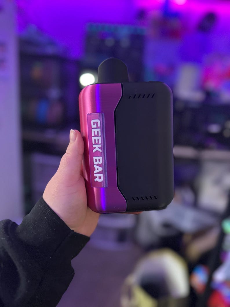 Peut inclure: Un appareil de vapotage Geek Bar. L'appareil a un corps noir avec un accent m&eacute;tallique violet. Le texte "GEEK BAR" est imprim&eacute; verticalement sur le c&ocirc;t&eacute; violet. L'appareil a un embout buccal noir.