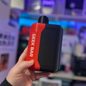 Peut inclure: Un appareil de vapotage Geek Bar noir et rouge. L'appareil porte l'inscription "GEEK BAR" en blanc sur un panneau rouge. L'appareil est rectangulaire avec un embout buccal noir et une s&eacute;rie de petits &eacute;vents.