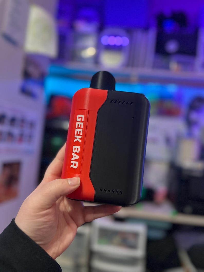 Peut inclure: Un appareil de vapotage Geek Bar noir et rouge. L'appareil porte l'inscription "GEEK BAR" en blanc sur un panneau rouge. L'appareil est rectangulaire avec un embout buccal noir et une s&eacute;rie de petits &eacute;vents.