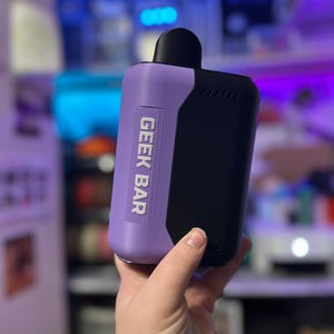 Peut inclure: Un appareil de vape Geek Bar bicolore. Le corps est lavande avec le texte "GEEK BAR" en blanc, et une section noire. L'embout buccal est noir. Tenu dans une main.