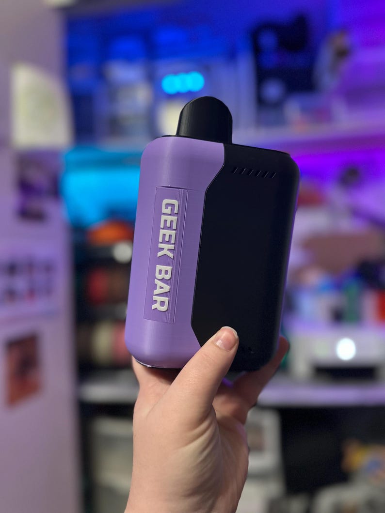 Peut inclure: Un appareil de vape Geek Bar bicolore. Le corps est lavande avec le texte "GEEK BAR" en blanc, et une section noire. L'embout buccal est noir. Tenu dans une main.