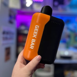 Könnte beinhalten: Ein orangefarbenes und schwarzes Geek Bar Vape-Gerät. Das Gerät hat eine rechteckige Form mit abgerundeten Kanten. Der Text "GEEK BAR" ist vertikal in Weiß auf dem orangefarbenen Abschnitt aufgedruckt. Die Oberseite ist schwarz und abgerundet.