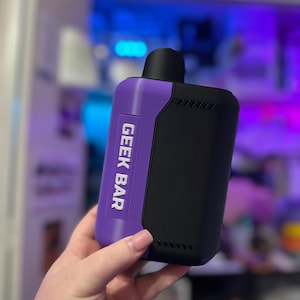 Peut inclure: Un appareil de vapotage Geek Bar noir et violet. L'appareil porte l'inscription "GEEK BAR" en blanc sur une section violette. L'appareil est rectangulaire avec un embout buccal noir et une s&eacute;rie de petits trous sur le c&ocirc;t&eacute;.