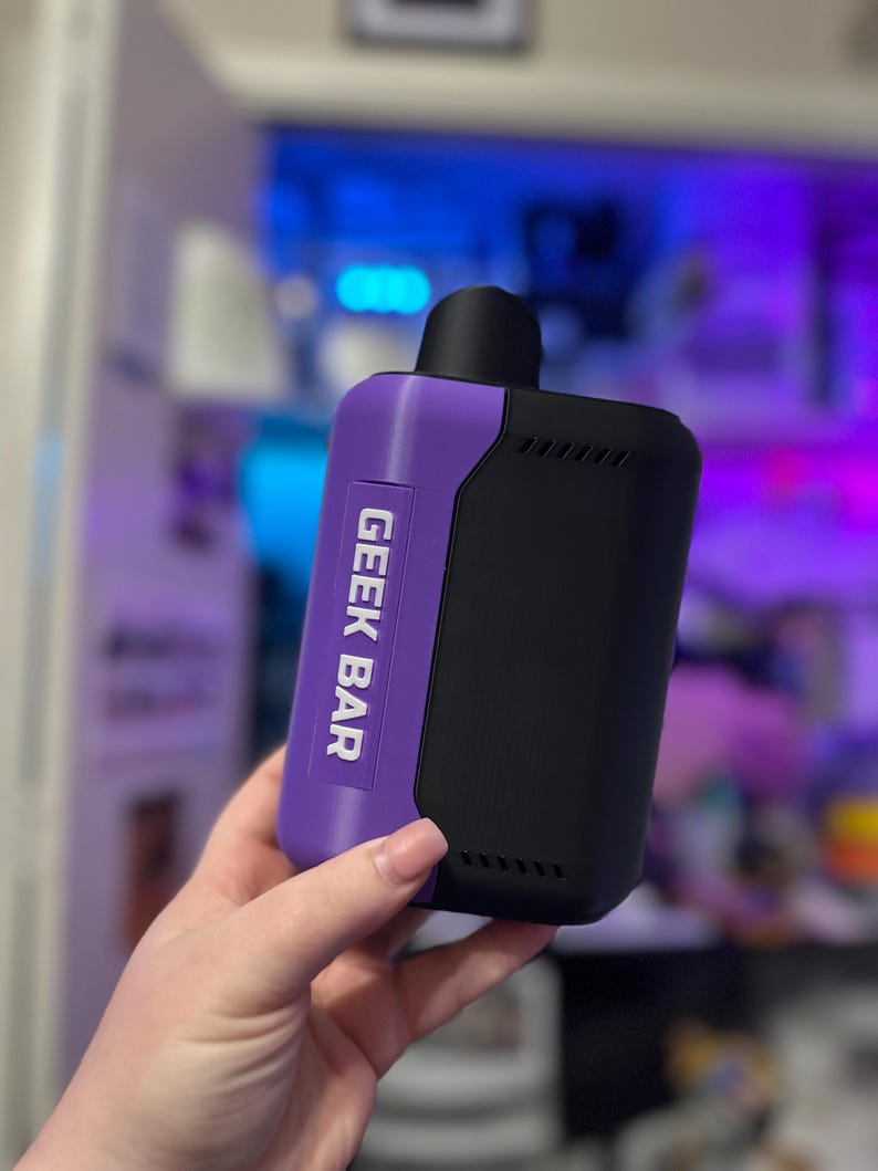 Peut inclure: Un appareil de vapotage Geek Bar noir et violet. L'appareil porte l'inscription "GEEK BAR" en blanc sur une section violette. L'appareil est rectangulaire avec un embout buccal noir et une s&eacute;rie de petits trous sur le c&ocirc;t&eacute;.