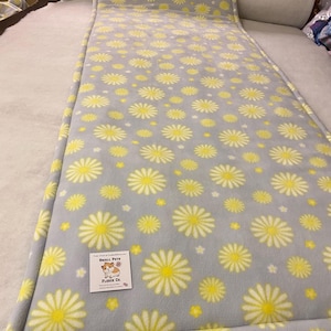 May include: A rectangular grey fleece pet mat featuring a repeating pattern of yellow and white daisy flowers. A small white square label is attached to the surface, displaying a hamster illustration and the text Small Pets Fleece Co.