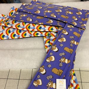 May include: A collection of rectangular fleece pads featuring two distinct patterns: one with a purple background decorated with brown and white guinea pigs and small white flowers, and another with a white background featuring colourful rainbows.