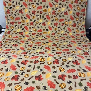 May include: A rectangular fleece blanket featuring a beige background with a repeating pattern of orange pumpkins, red and yellow autumn leaves, small black squirrels, and brown twigs. The fabric has a soft, textured surface with finished edges.