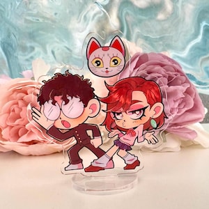 Puede incluir: Un soporte acrílico transparente con dos figuras de dibujos animados y una cabeza de gato. Las figuras son un niño con un traje marrón y una niña con una camisa rosa y una falda morada. La cabeza del gato es rosa y blanca.