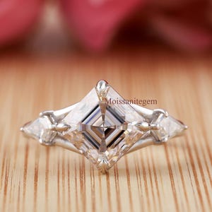 May include: A silver ring with a large, square-cut, clear gemstone as the centerpiece, flanked by two smaller triangular stones. The ring is set on a wooden surface, with the text "Moissanitegem" visible.
