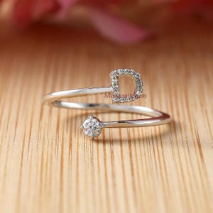 Puede incluir: Un anillo plateado con un diseño abierto único. El anillo presenta una piedra preciosa redonda y brillante y una forma de letra "D" incrustada con piedras más pequeñas. El anillo está sobre una superficie de madera cálida.