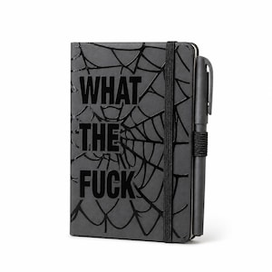 May include: A black notebook with a glossy spider web pattern embossed on the cover. The text WHAT THE FUCK is printed in large black capital letters on the front. A black elastic band holds the notebook closed, and a black pen is attached to the side.