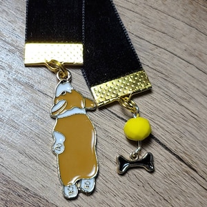 May include: Black velvet bookmark with a gold metal charm of a brown and white corgi dog with a yellow bead and a black bone charm.