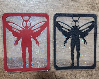 May include: Two clear plastic frames with a glittery background. Each frame contains a silhouette of a winged creature with large eyes. One creature is red and the other is black.