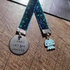 May include: A teal blue glitter bookmark with a silver charm that says "I shall call you squishy" and a blue and silver jellyfish charm.