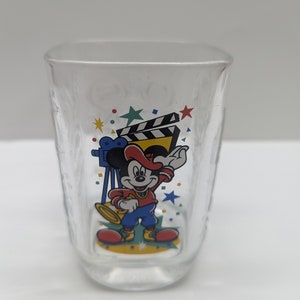 Puede incluir: Una taza de vidrio transparente con un diseño colorido que presenta a Mickey Mouse vestido de director de orquesta, sosteniendo una trompeta, con una claqueta de cine y una cámara de cine en el fondo. El diseño incluye estrellas y confeti.