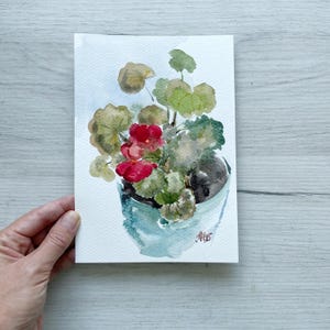 May include: Watercolor painting of a potted geranium plant with red flowers and green leaves. The plant is in a light blue pot. The artwork is on white paper.