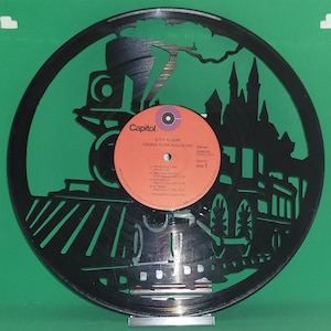 May include: A black vinyl record with a silhouette of a train engine and a cityscape. The record label is red and white with the text "Capitol. LIVE ALBUM GRAND FUNK RAILROAD Stereo SWB 11231  Side 1" printed on it.