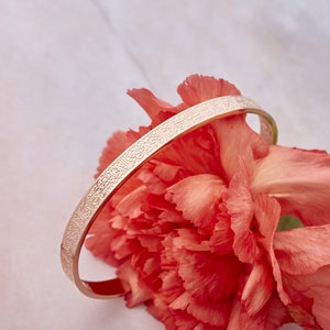 May include: A rose gold bangle bracelet with an intricate engraved design. The bracelet is resting on a pink flower.