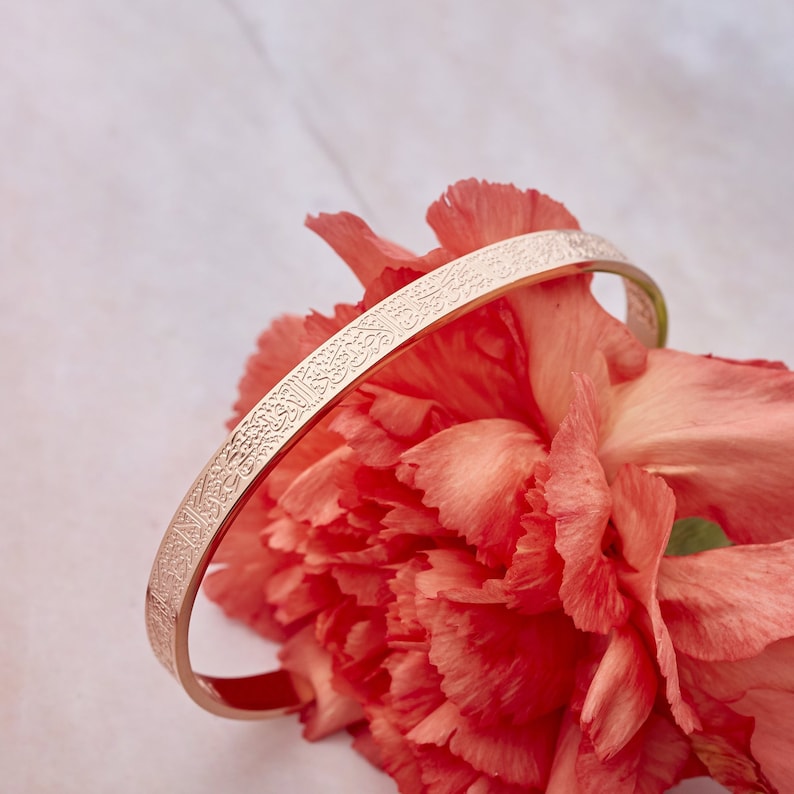 May include: A rose gold bangle bracelet with an intricate engraved design. The bracelet is resting on a pink flower.