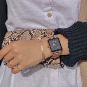 May include: A rose gold square-shaped watch with a black face and a mesh band. The watch is on a person's wrist.