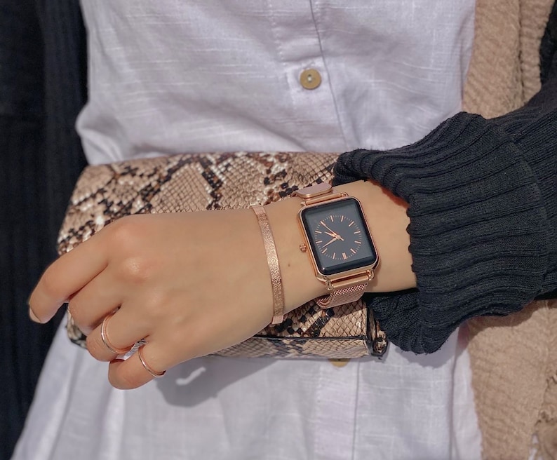 May include: A rose gold square-shaped watch with a black face and a mesh band. The watch is on a person's wrist.