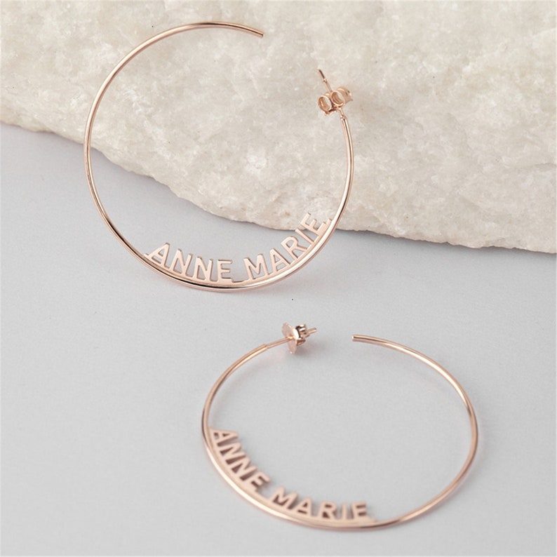 May include: A pair of rose gold hoop earrings with the name "Anne Marie" cut out of the metal.