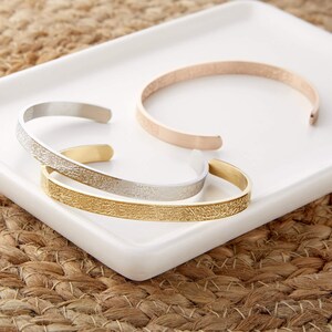 May include: Three gold, silver, and rose gold bracelets with an intricate engraved pattern. The bracelets are stacked on a white surface.