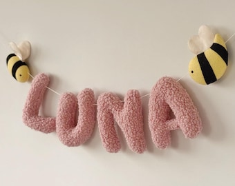 May include: A pink, plush banner spelling "LUNA" is strung between two fabric bees. The bees are yellow and black with white wings. The banner is designed for a nursery or child's room.
