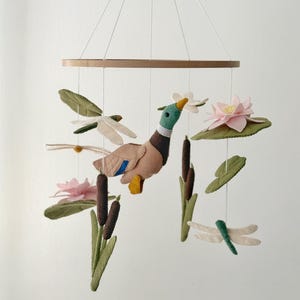 May include: A handcrafted baby mobile featuring a duck, dragonflies, lily pads, and cattails. The duck has a green head, brown body, and yellow feet. The mobile is suspended from a wooden ring with white strings. The color palette includes shades of green, pink, and brown.