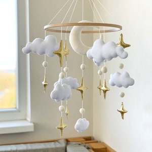 Op de afbeelding: Een witte en gouden babymobiel van vilt met wolken, sterren en een halve maan die aan een houten ring hangt.