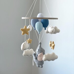 May include: A baby mobile featuring a gray elephant holding onto blue balloons, with white clouds and gold stars. The mobile is suspended from a wooden ring and is designed for a nursery.