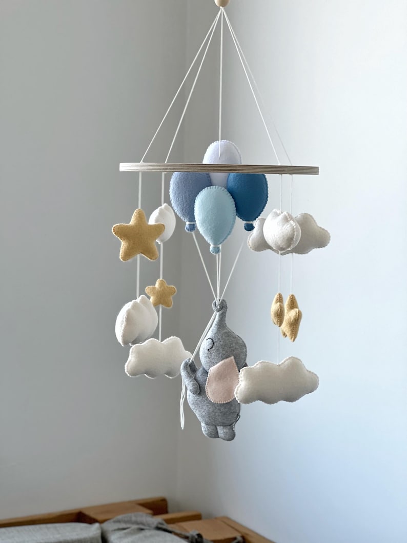 May include: A baby mobile featuring a gray elephant holding onto blue balloons, with white clouds and gold stars. The mobile is suspended from a wooden ring and is designed for a nursery.