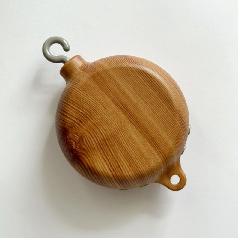 May include: A round, wooden object with a light brown finish and a gray metal hook. The wood grain is visible, and the object has a small hole on the side. The hook is attached to the top.