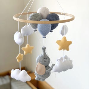 May include: A baby mobile featuring a wooden hoop with felt balloons, clouds, stars, and an elephant. The balloons are in shades of gray and blue, while the stars are yellow. The elephant has a beige bib.