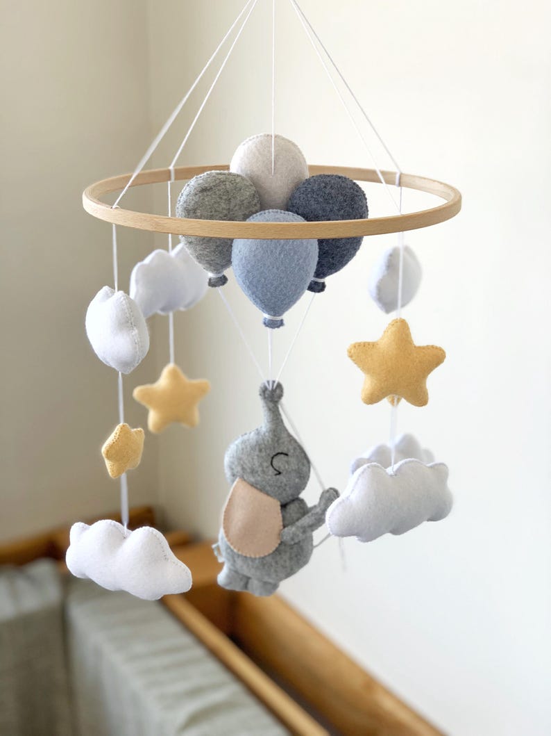 May include: A baby mobile featuring a wooden hoop with felt balloons, clouds, stars, and an elephant. The balloons are in shades of gray and blue, while the stars are yellow. The elephant has a beige bib.
