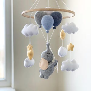 May include: A baby mobile featuring a gray felt elephant holding onto balloons. The mobile includes white clouds, yellow stars, and gray and blue balloons suspended from a wooden hoop. The elephant has a light pink ear.