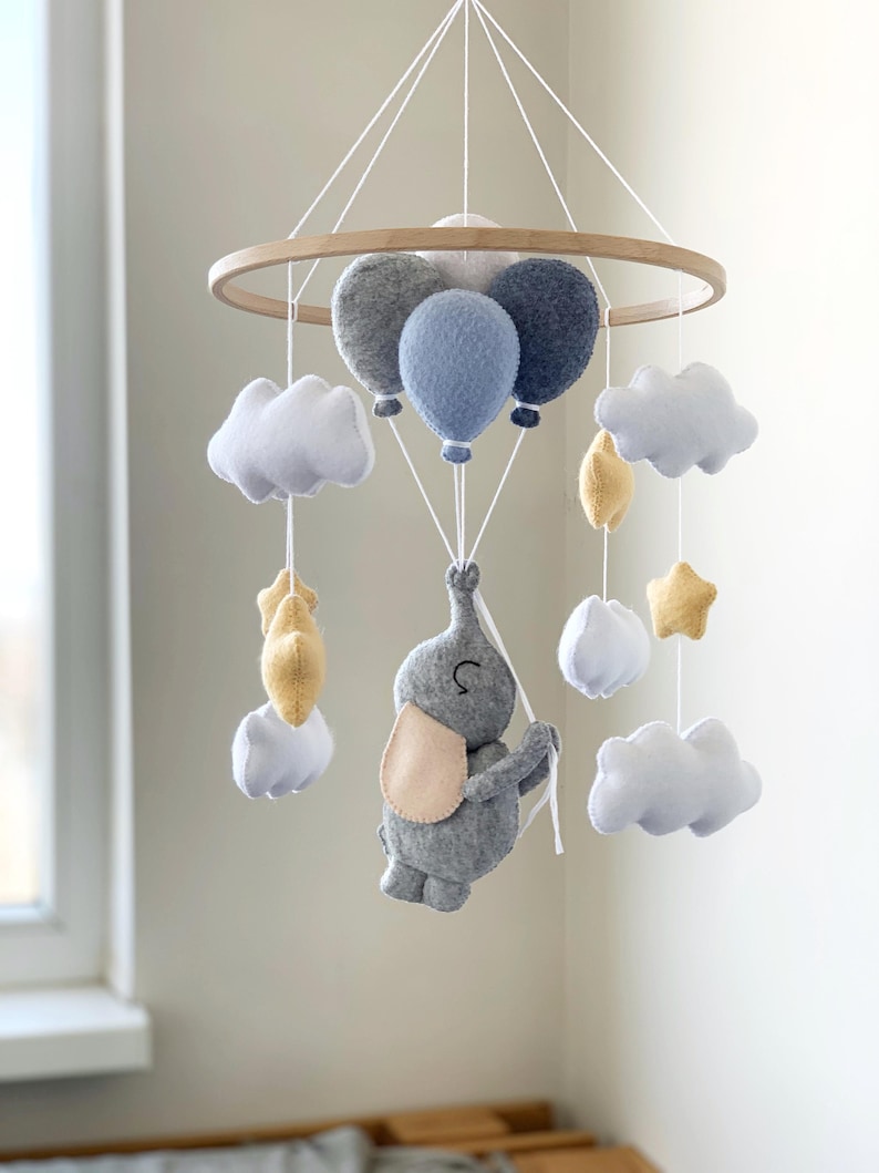 May include: A baby mobile featuring a gray felt elephant holding onto balloons. The mobile includes white clouds, yellow stars, and gray and blue balloons suspended from a wooden hoop. The elephant has a light pink ear.