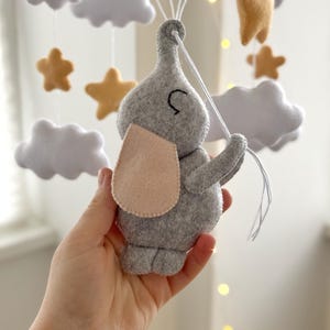 May include: A gray felt elephant mobile with a light pink ear, holding strings attached to blue felt clouds. The mobile also features white felt clouds and gold felt stars. A whimsical nursery decoration.
