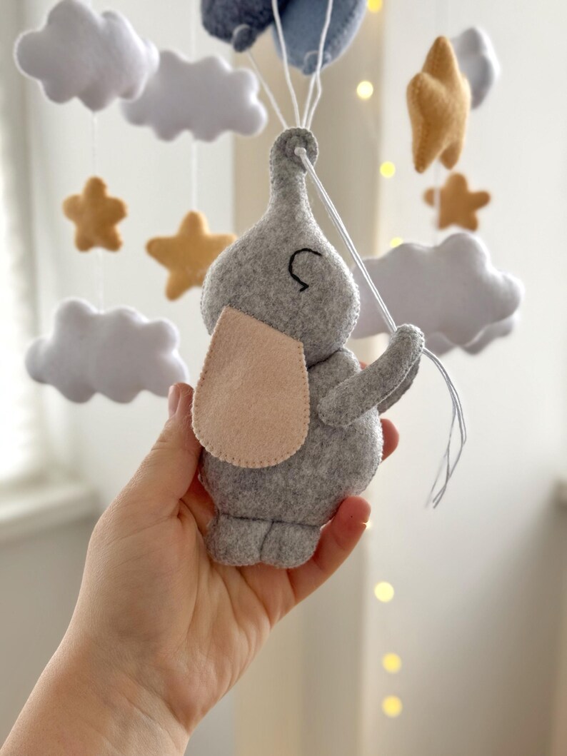 May include: A gray felt elephant mobile with a light pink ear, holding strings attached to blue felt clouds. The mobile also features white felt clouds and gold felt stars. A whimsical nursery decoration.