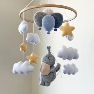 May include: A baby mobile featuring a wooden hoop with hanging felt shapes. The mobile includes gray and blue balloons, white clouds, yellow stars, and a gray elephant with a pink ear. The text "Option 2" is visible.