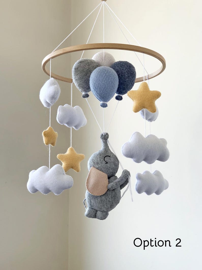 May include: A baby mobile featuring a wooden hoop with hanging felt shapes. The mobile includes gray and blue balloons, white clouds, yellow stars, and a gray elephant with a pink ear. The text "Option 2" is visible.