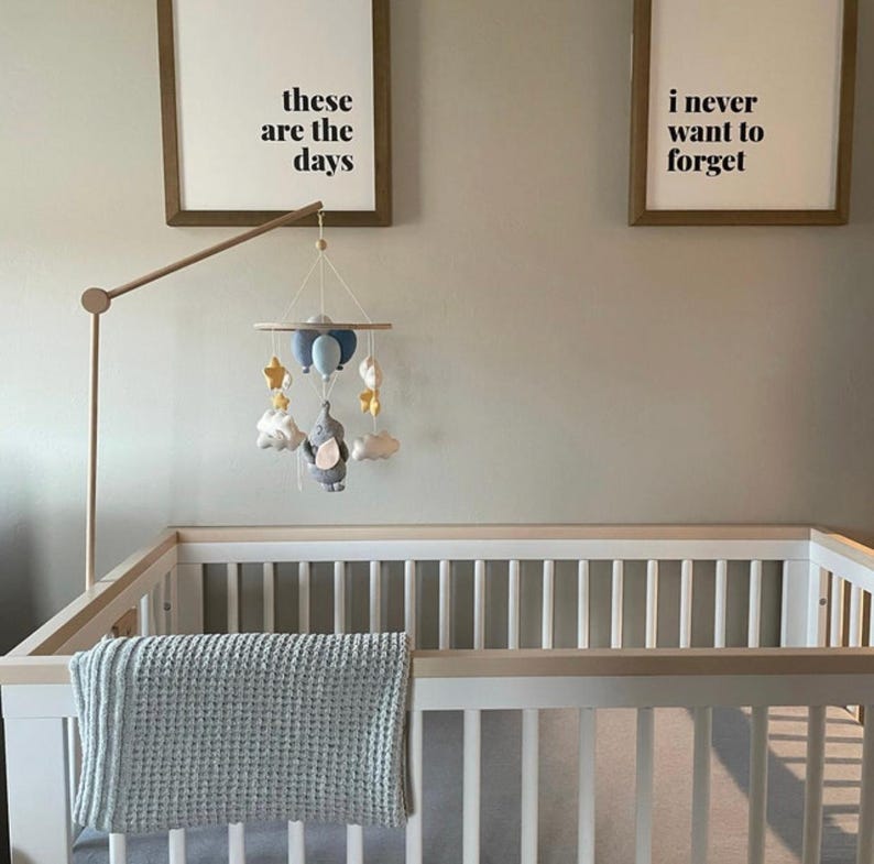 May include: A baby crib with a light blue blanket and a mobile featuring clouds, stars, and an elephant. Two framed prints above the crib read "these are the days" and "i never want to forget".