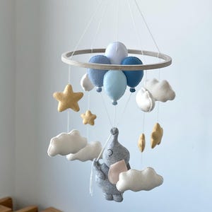 May include: A baby mobile featuring a gray elephant holding balloons and surrounded by clouds and stars. The mobile has a wooden ring and is suspended by strings. The balloons are various shades of blue, and the stars are gold.