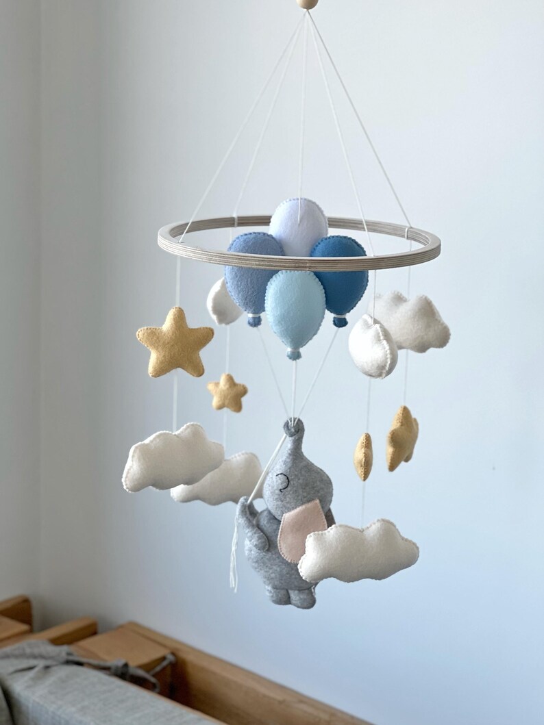 May include: A baby mobile featuring a gray elephant holding balloons and surrounded by clouds and stars. The mobile has a wooden ring and is suspended by strings. The balloons are various shades of blue, and the stars are gold.