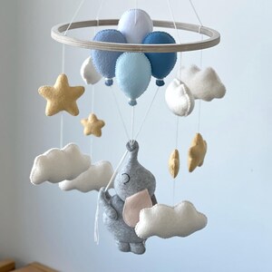 May include: A baby mobile featuring a gray elephant with a pink ear, suspended below blue balloons, white clouds, and gold stars. The mobile is attached to a wooden ring with white strings. The text "Option 1" is visible.