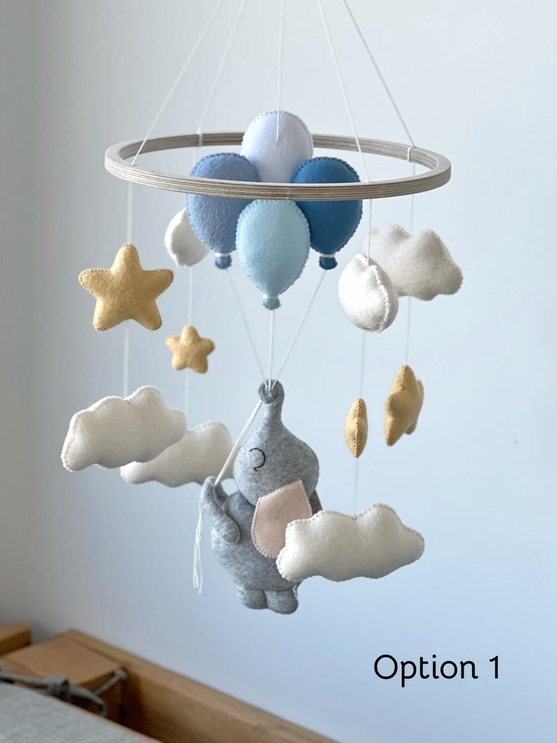 May include: A baby mobile featuring a gray elephant with a pink ear, suspended below blue balloons, white clouds, and gold stars. The mobile is attached to a wooden ring with white strings. The text "Option 1" is visible.