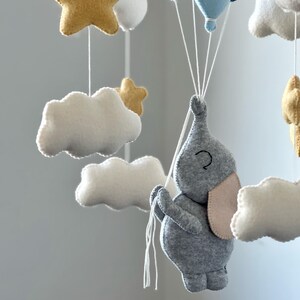 May include: A handmade baby mobile featuring a gray felt elephant holding blue felt balloons. The mobile also includes white felt clouds and gold felt stars, all suspended by white strings. A charming nursery decoration.