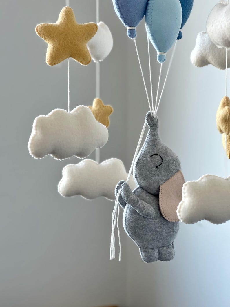 May include: A handmade baby mobile featuring a gray felt elephant holding blue felt balloons. The mobile also includes white felt clouds and gold felt stars, all suspended by white strings. A charming nursery decoration.