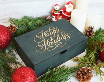 May include: A dark green wooden box with the words "Happy Holidays" in gold script. The box is surrounded by evergreen branches, red glitter baubles, pine cones, and white candles. Festive holiday decor.