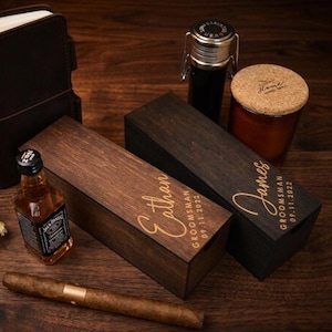 May include: A collection of gifts for a special occasion, including a leather-bound journal, a bottle of liquor, a cigar, and two wooden boxes engraved with names and dates. A candle in a glass jar is also present.